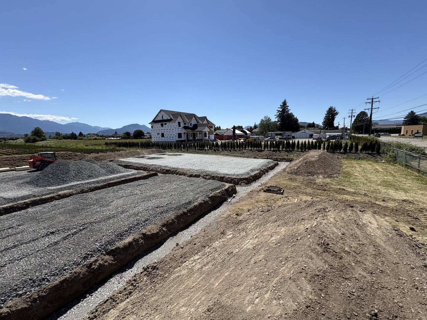 A gravel building pad and a cut drainage trench on a cleared lot, a new house under construction behind